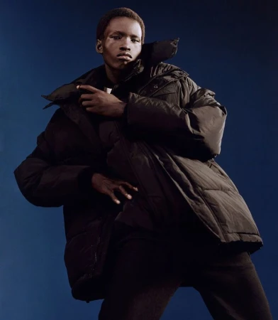Dark-skinned adult fashion model poses in an oversized, dark puffer jacket and matching pants against a dark blue studio backdrop.