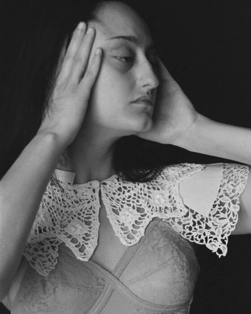 Monochrome portrait of a young woman with hands framing her face, wearing a lace collar and bra, set against a dark background.