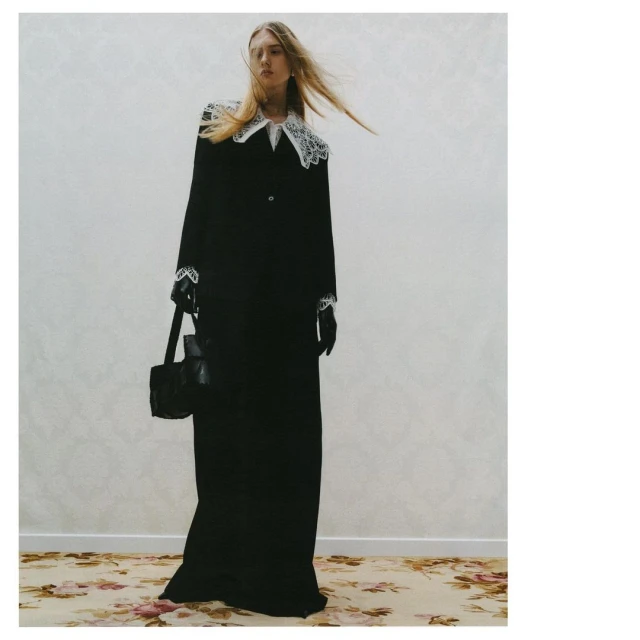 A fashionable woman in a black gown with white lace details poses indoors with a black handbag on a floral rug and neutral wall.