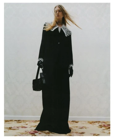 A fashionable woman in a black gown with white lace details poses indoors with a black handbag on a floral rug and neutral wall.