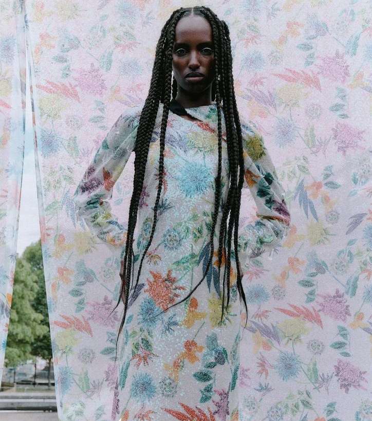 A young Black woman poses wearing a floral dress with an outdoor backdrop, her hair styled in long braids. Fashion editorial portrait.