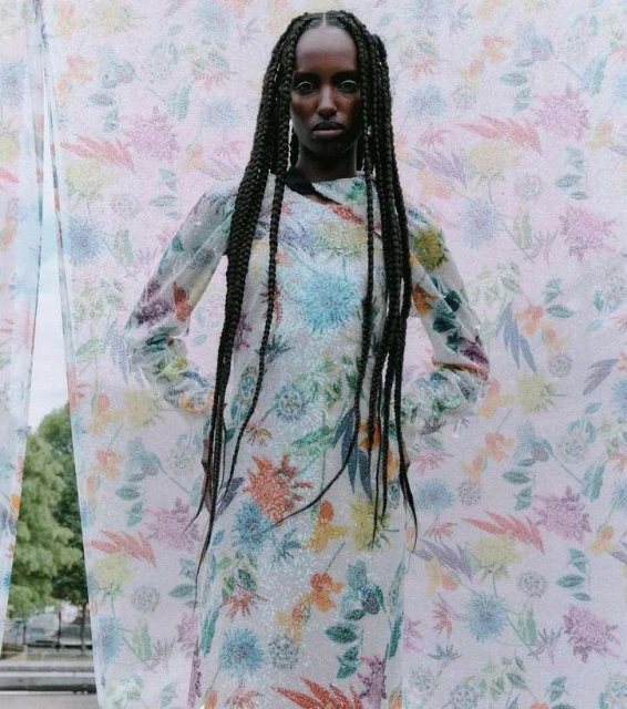 A young Black woman poses wearing a floral dress with an outdoor backdrop, her hair styled in long braids. Fashion editorial portrait.