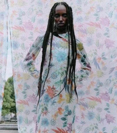 A young Black woman poses wearing a floral dress with an outdoor backdrop, her hair styled in long braids. Fashion editorial portrait.