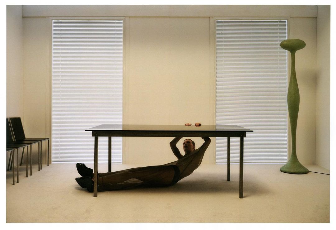 Adult under a dark table in a bright interior with blinds, chairs, a green floor lamp and beige walls, creating a modern, minimalist scene.