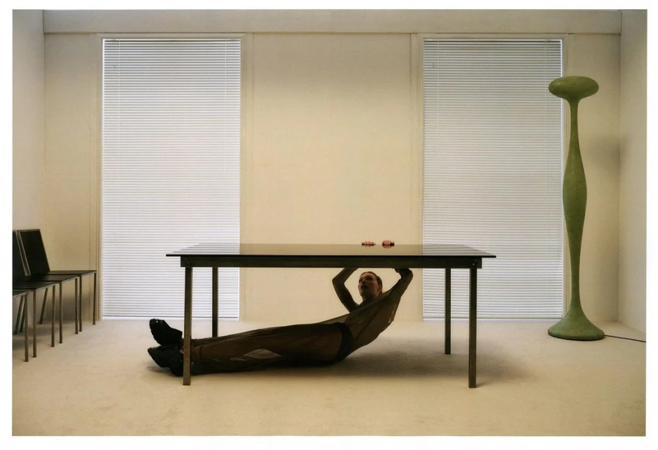 Adult under a dark table in a bright interior with blinds, chairs, a green floor lamp and beige walls, creating a modern, minimalist scene.