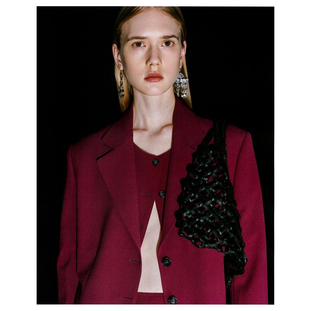 Blonde woman in burgundy blazer and top with black woven bag against a black background in a studio portrait with soft lighting.