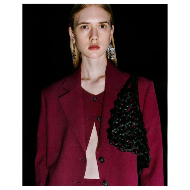 Blonde woman in burgundy blazer and top with black woven bag against a black background in a studio portrait with soft lighting.