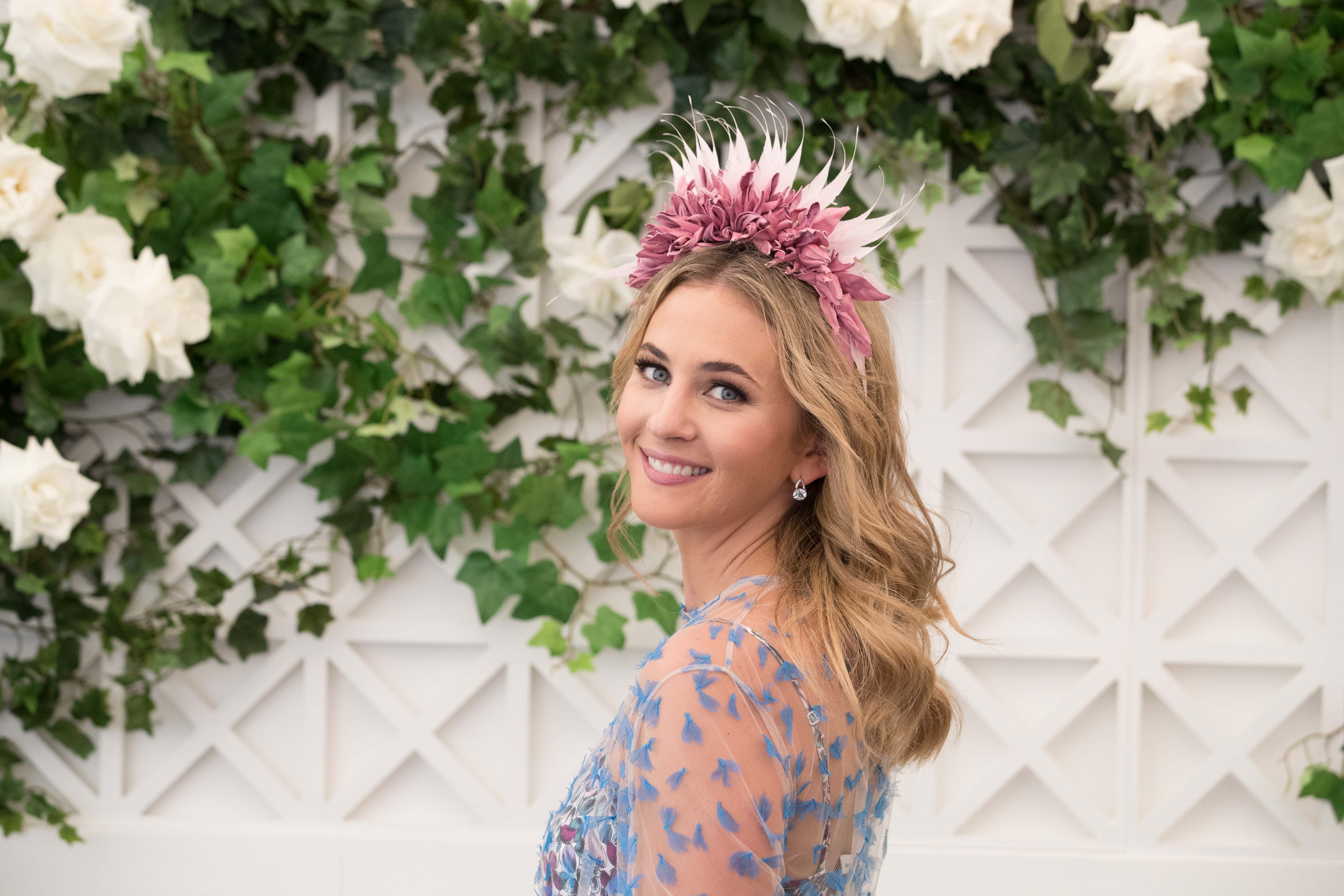 Smiling woman with blonde wavy hair wearing a mauve floral headpiece, floral top, standing in front of white roses and leafy green vines.