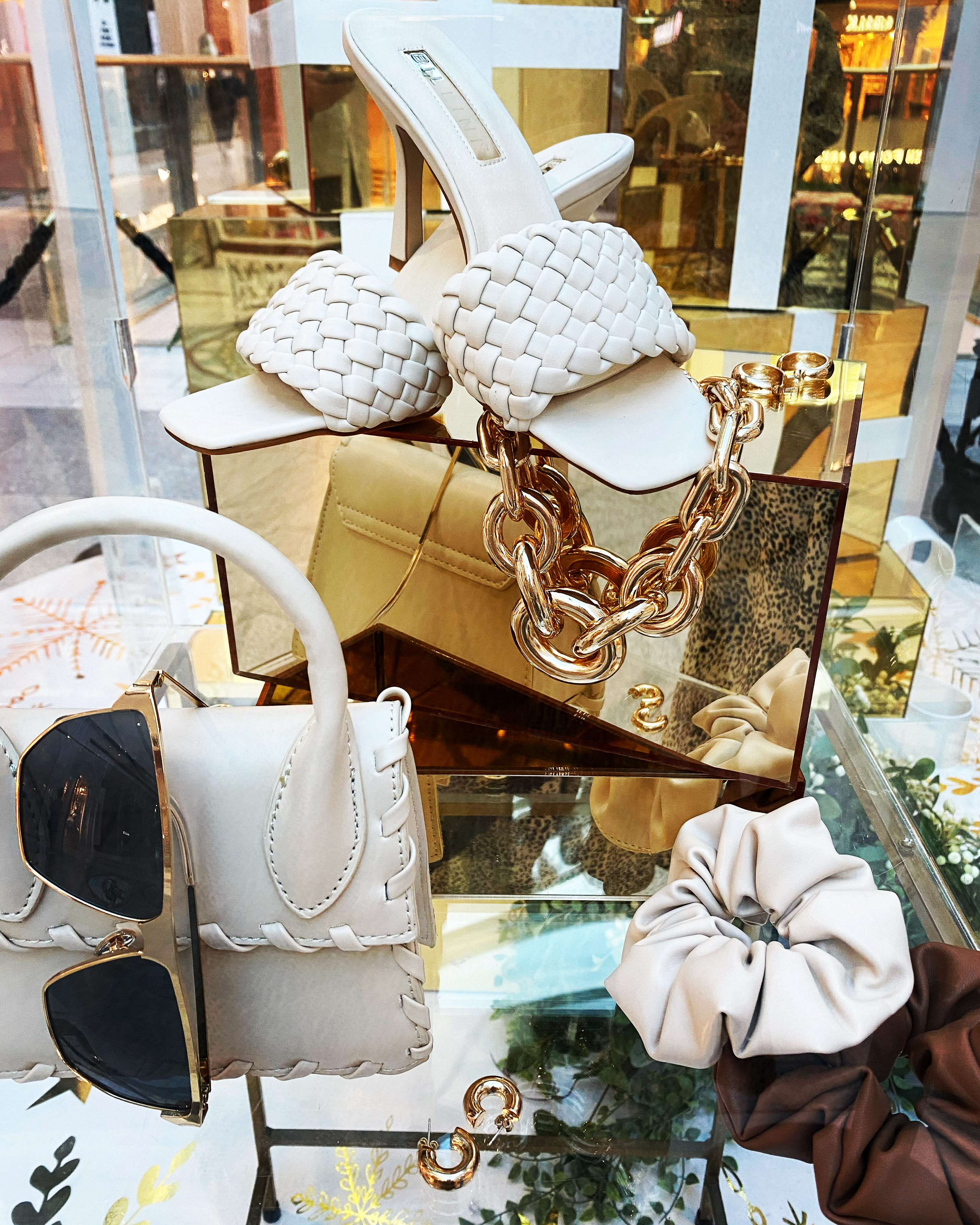 High-angle shot of curated women's fashion accessories including woven heeled sandals, gold purse with chain, handbag, earrings, and scrunchies.