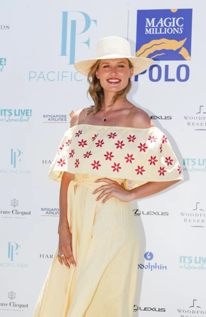 Blonde woman in off-shoulder dress and hat poses at Pacific Polo event with branded signage backdrop and logos. Outdoor, daytime.