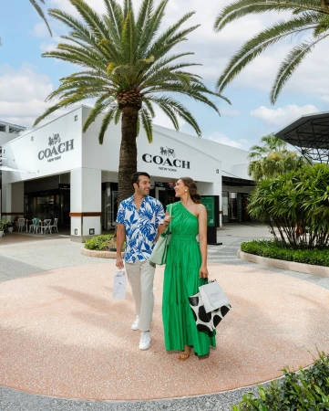 A stylish couple walks past a Coach store, carrying shopping bags, on a sunny day with palm trees in a bright, outdoor shopping center.