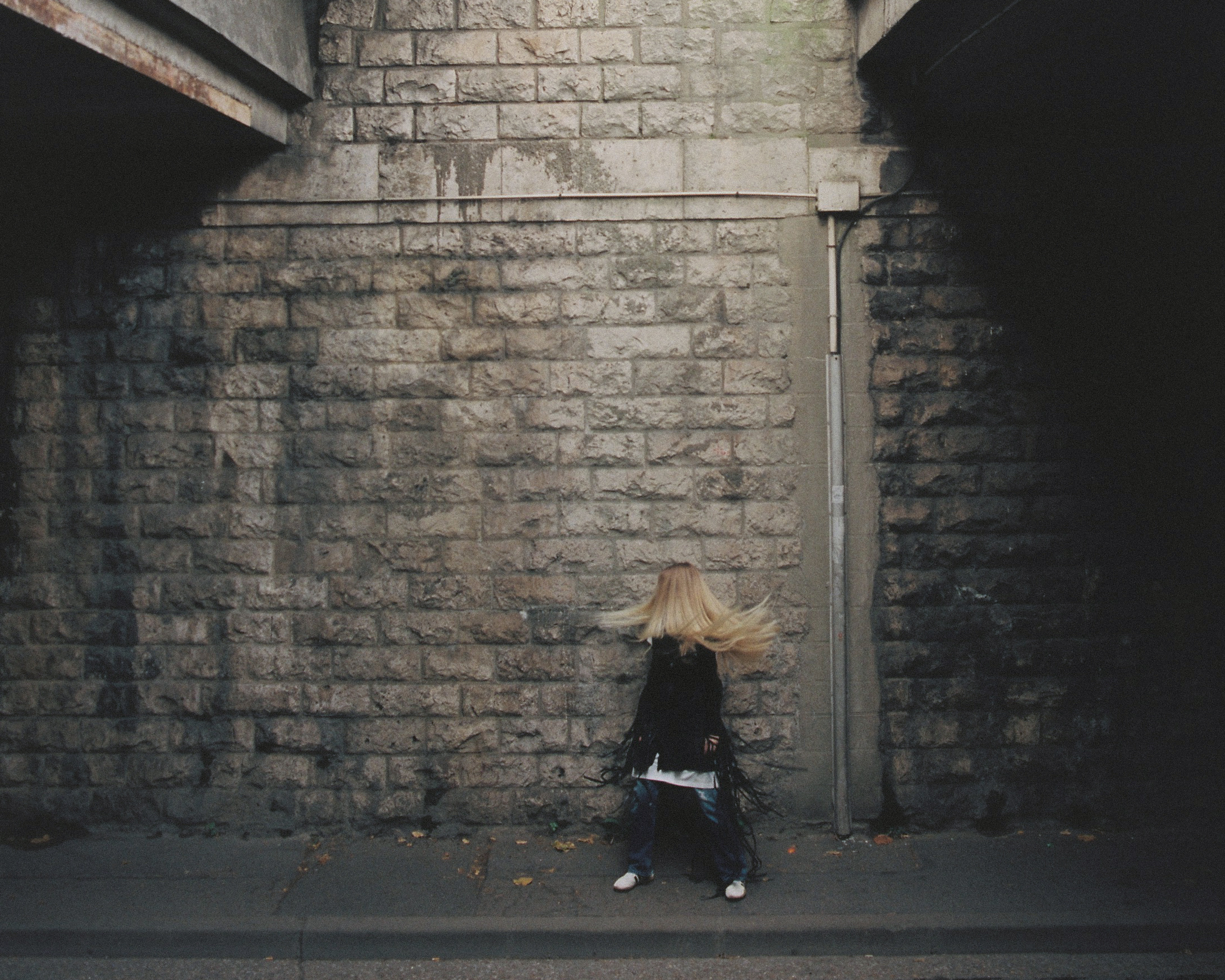 Blond woman dances in front of a stone wall under a concrete bridge, wearing a fringed black jacket, jeans, and white shoes.