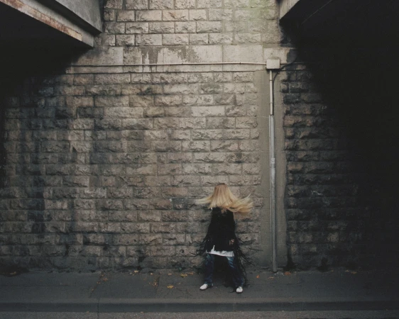 Blond woman dances in front of a stone wall under a concrete bridge, wearing a fringed black jacket, jeans, and white shoes.