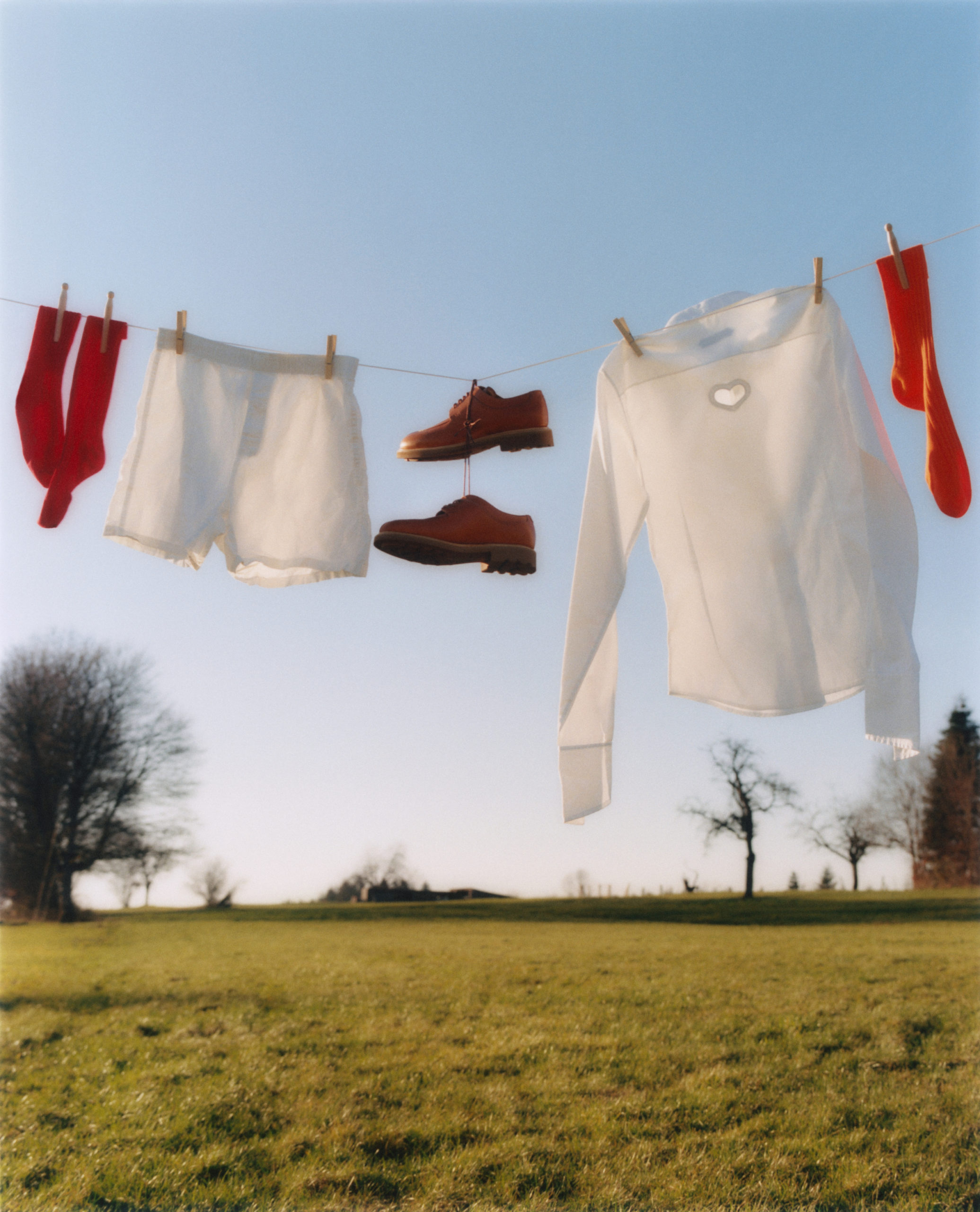 Outdoor clothesline with socks, shirt, shorts, and shoes held by clothespins against a blue sky over a green field and trees.