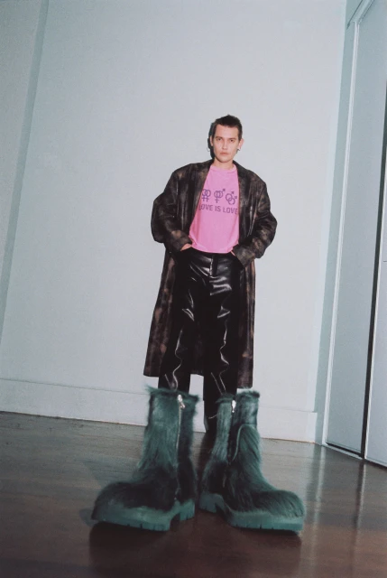 An adult stands indoors wearing a pink t-shirt, leather pants, a trench coat, and furry boots, posing in front of a white wall.