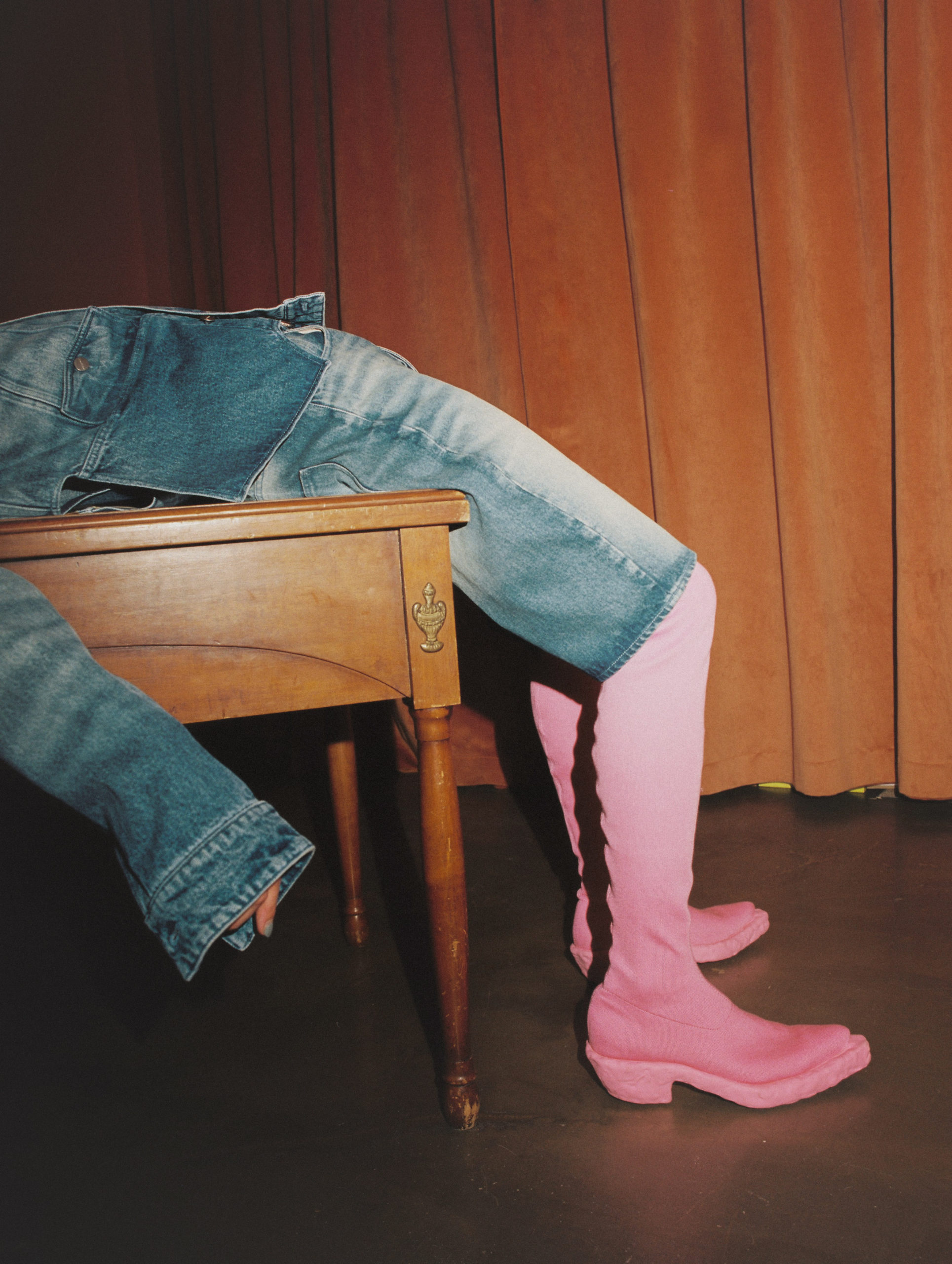 Person wearing pink boots and denim jacket and jeans, reclining on a wooden desk in front of a brown curtain in a studio.