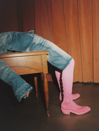 Person wearing pink boots and denim jacket and jeans, reclining on a wooden desk in front of a brown curtain in a studio.