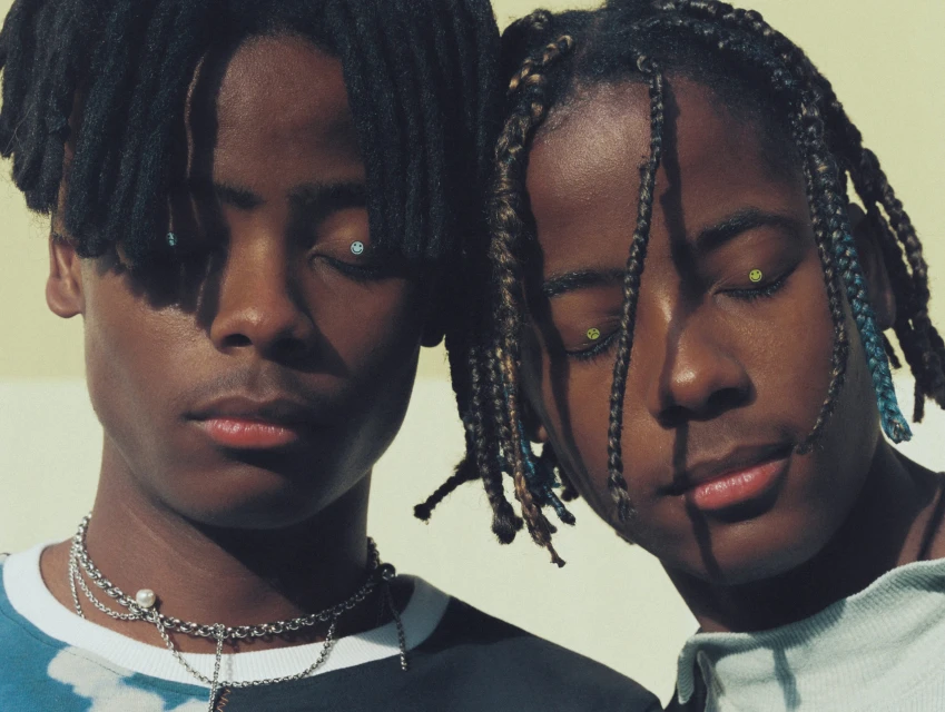 Close-up portrait of two dark-skinned young adult males with unique hairstyles posing indoors with jewelry and casual clothing.