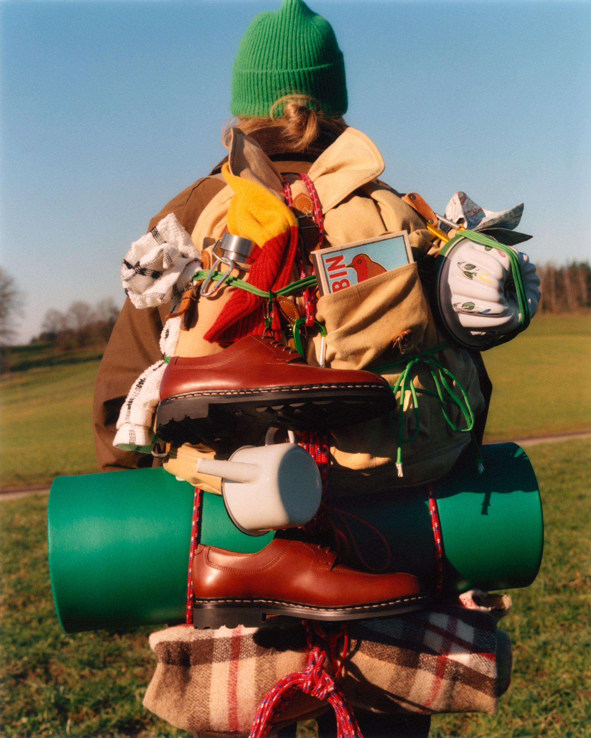 Backpack and hiking equipment, including boots, green sleeping bag, rope, plaid blanket, and a mug, against a sunny rural background.