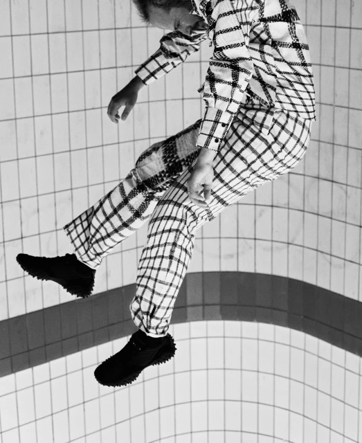 Monochromatic shot of a slender person mid-air in a matching checkered shirt and pants with black shoes, against tiled backdrop.