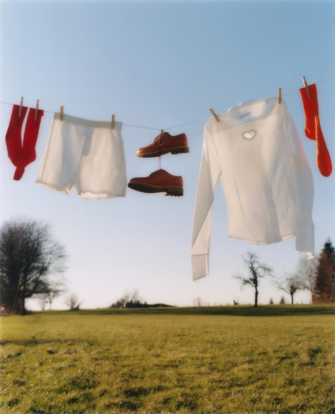 White garments, red socks, and brown shoes hanging on a clothesline outdoors against a grassy field and clear blue sky.