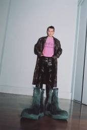 Model wearing oversized green furry boots, pink graphic t-shirt, and leather coat standing in a room with white walls.