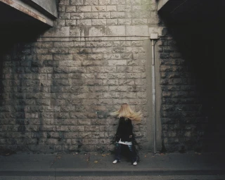 Woman with long blonde hair tossed across her face standing against a textured stone wall under a concrete bridge.