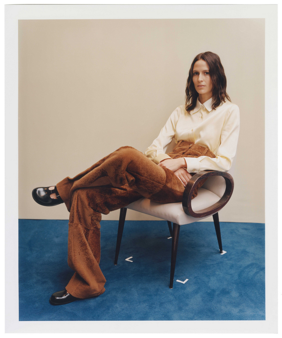 Woman in casual collared shirt and corduroy trousers sitting in a chair on a blue carpet in a photo studio with neutral backdrop.
