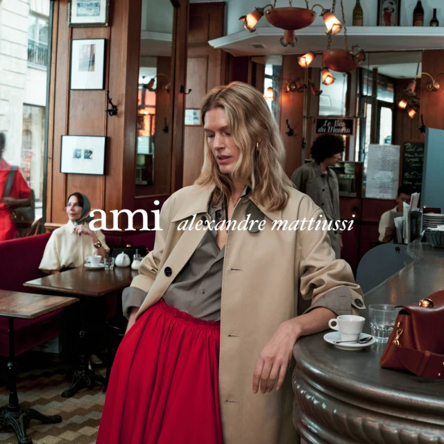Indoor fashion shot of a woman in a trench coat and red skirt standing at a counter with a coffee cup in a cafe setting.