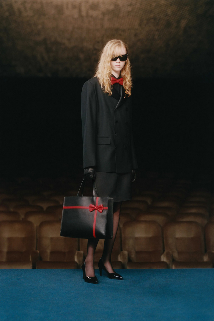 Adult woman poses in a dark theatre holding a black leather handbag with a red ribbon. She wears a black suit, and sunglasses.