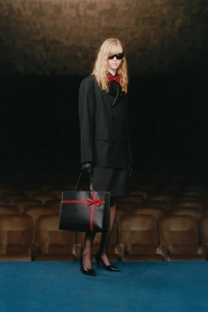 Adult woman poses in a dark theatre holding a black leather handbag with a red ribbon. She wears a black suit, and sunglasses.