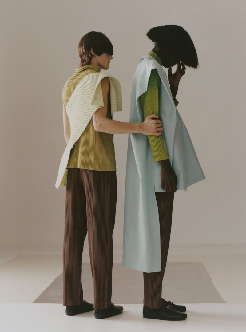 Two fashion models stand together in a studio setting, wearing stylish outfits in earth tones with modern, structured designs and black shoes.