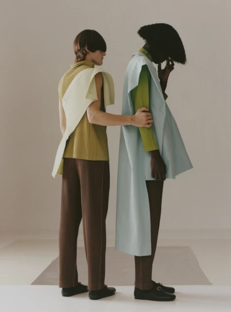Two fashion models stand together in a studio setting, wearing stylish outfits in earth tones with modern, structured designs and black shoes.