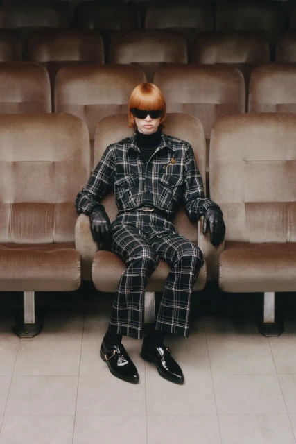 Stylish person with bowl cut and sunglasses in plaid outfit sits in empty cinema seat, showcasing retro fashion in cinematic setting.