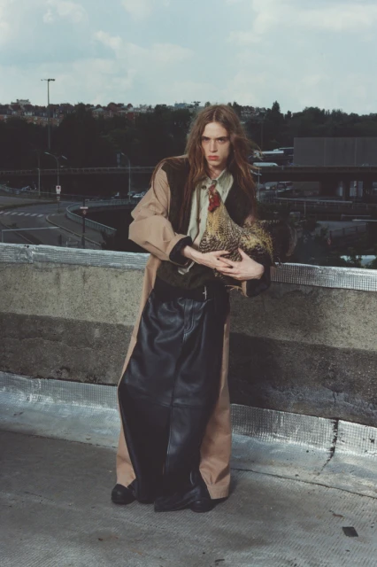 Adult male holding a rooster outdoors in an urban setting. He wears a long coat and leather pants with a city background.