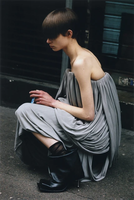 A woman with a bowl haircut squats on a city street wearing a grey dress and black boots. She is holding a blue object in her hands.