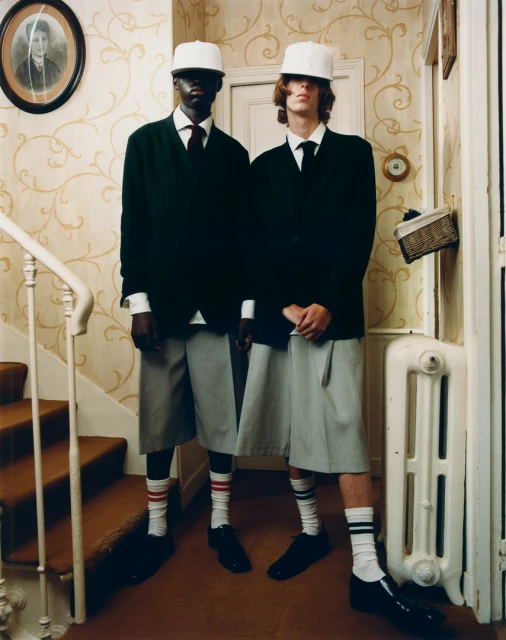 Two young men in matching dark blazers, light shorts, white socks with stripes, and white caps stand indoors against patterned wallpaper.