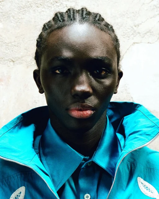 Close-up portrait of a young adult Black male with braided hair, wearing a turquoise jacket and collared shirt, against a neutral background.