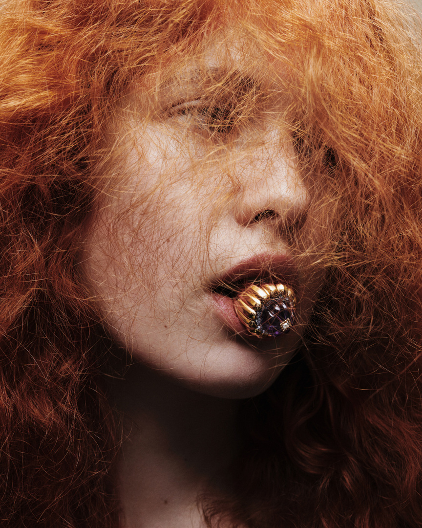 Close-up portrait of a woman with red hair holding a large gold ring in her mouth with hair falling across her face.