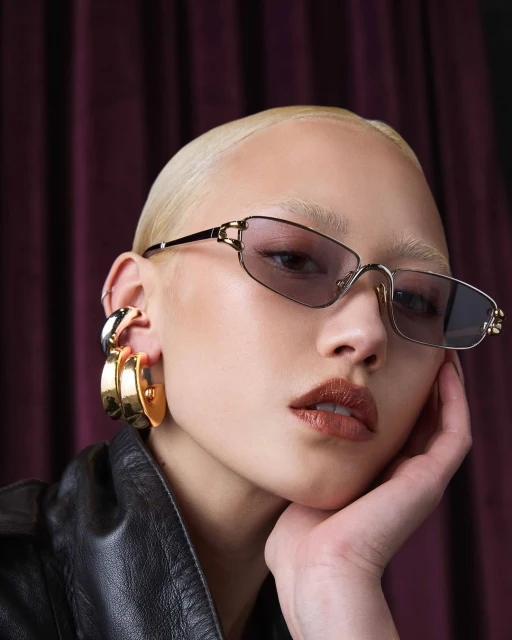 Close-up portrait of a stylish adult female model with pale skin and a shaved head wearing sunglasses, large earrings, and a black jacket.