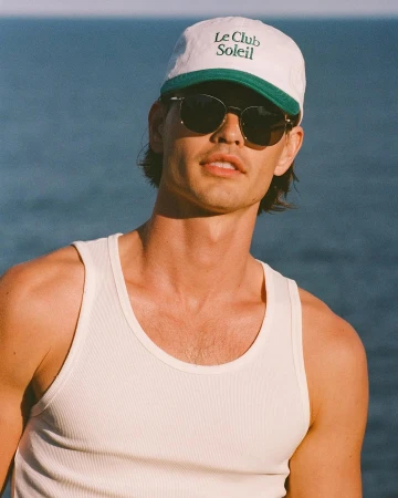 Man wearing a white and green baseball cap and dark sunglasses with a white ribbed tank top against an ocean background