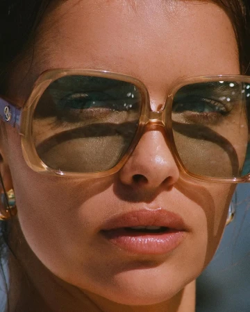 Close-up portrait of a woman wearing oversized tinted sunglasses with a warm golden hue and glossy lips.