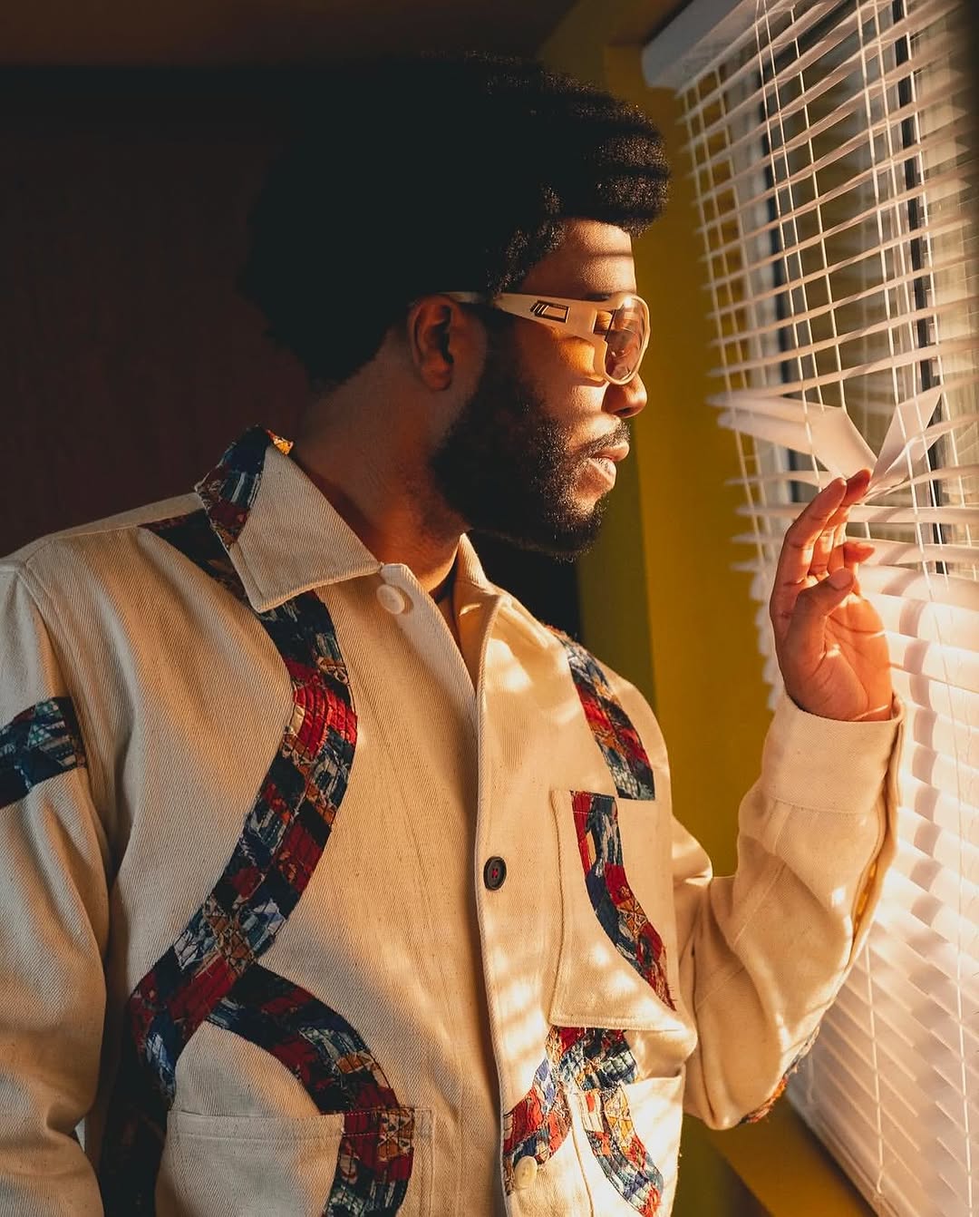 Stylish adult male in sunglasses and a patterned jacket looking out a window with blinds, bathed in warm, directional light.