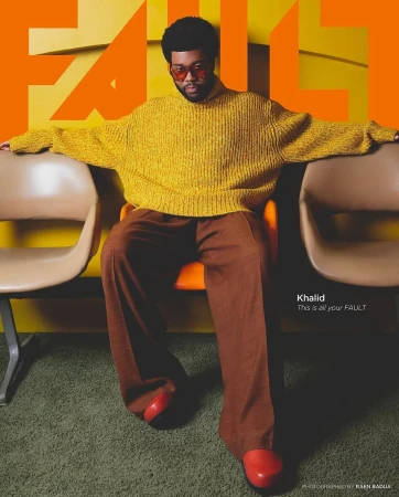 Khalid seated between two chairs wearing a yellow chunky knit sweater and brown trousers against yellow.