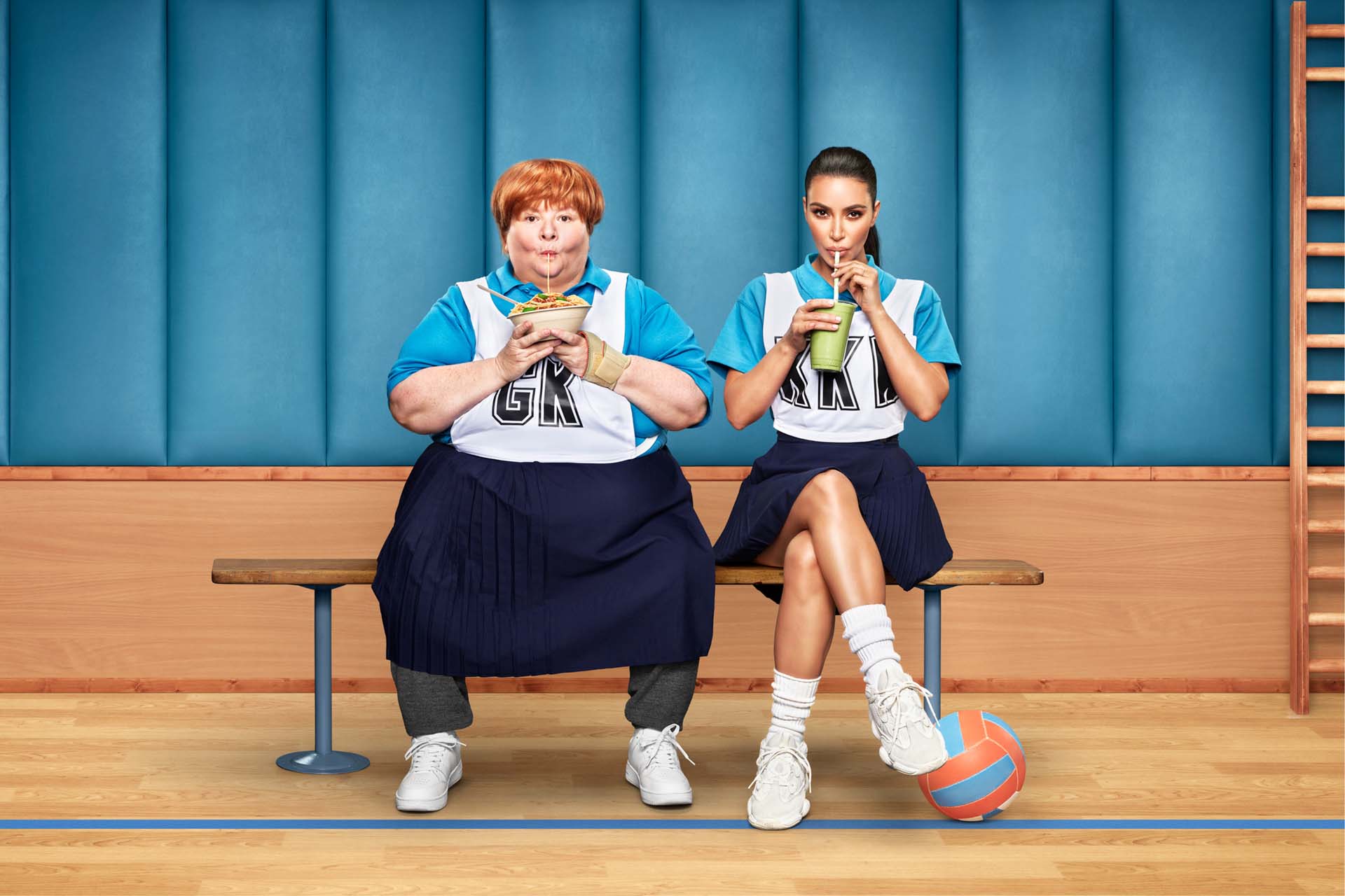 Two women in sports uniforms sit side-by-side on a bench indoors. One drinks a green smoothie, the other eats noodles from a bowl.