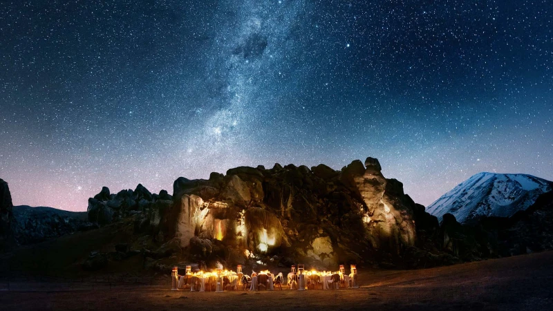 Outdoor dining tables are set with plates and glassware in front of a rocky cliff face at night beneath a sky full of stars.