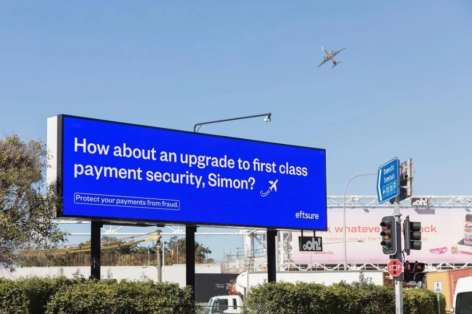 Outdoor advertising billboard with airline and payment security message against clear blue sky with a plane flying above.