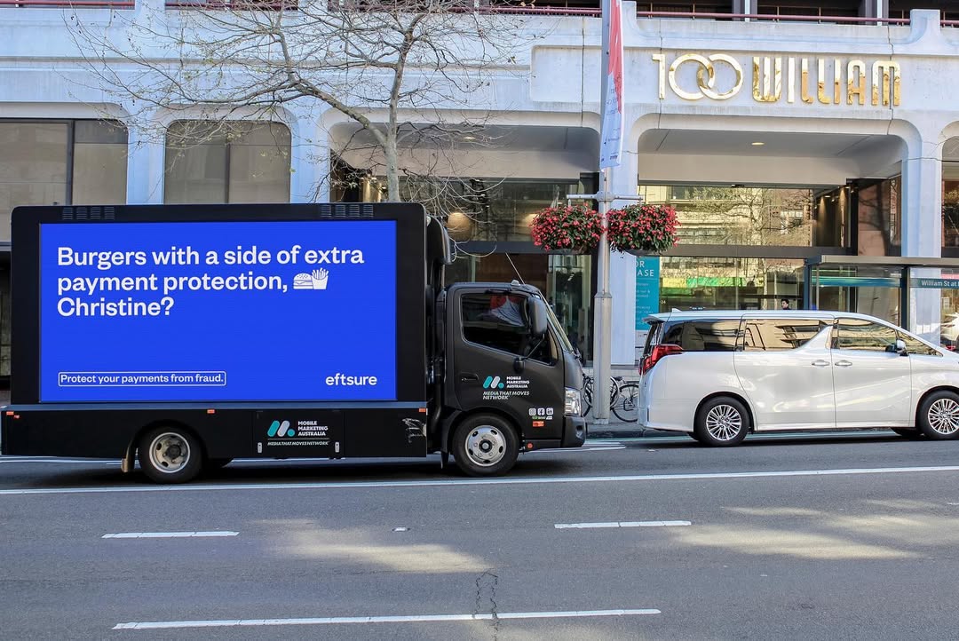 A mobile advertising truck drives on a city street with a digital ad for a payment protection service featuring a burger and fries.