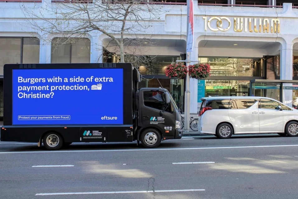 A mobile advertising truck drives on a city street with a digital ad for a payment protection service featuring a burger and fries.