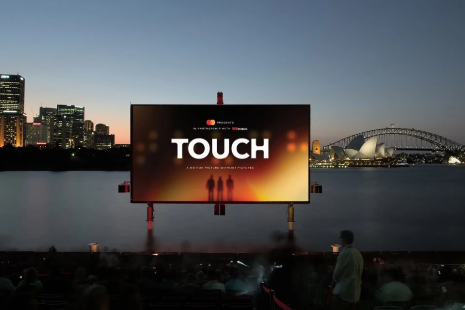Outdoor film screening at dusk featuring a large screen displaying the word 'TOUCH' with the Sydney Opera House in the background.